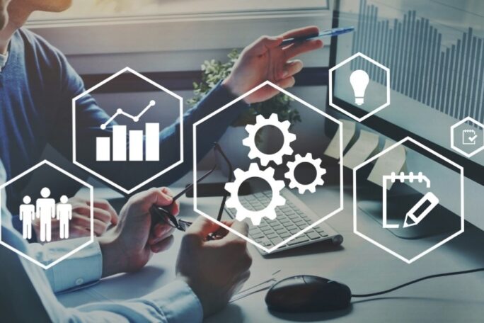 Five hexagons that have imagery inside them, including people, bar charts, gears, light bulbs, and a pad of paper. Behind the hexagons are two people reviewing data on a computer screen